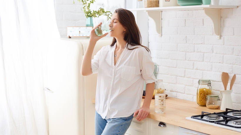 A woman with brown hair and wearing a white shirt with jeans is drinking water in her kitchen the day of her tennis match.