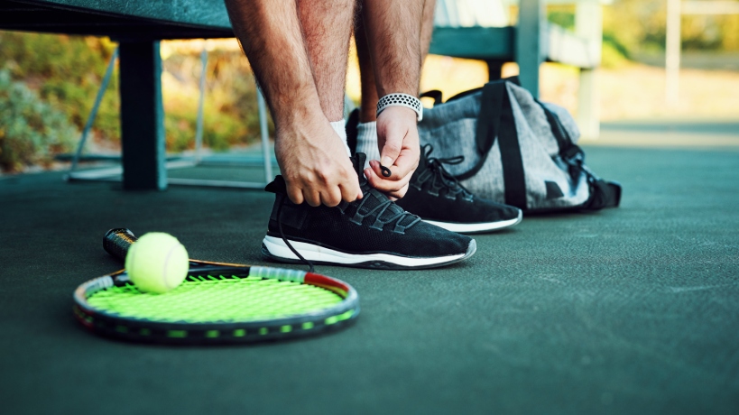 A man wearing a white bracelet lacing up his black tennis shoes with a tennis racket and ball to his right and his tennis bag to his left.