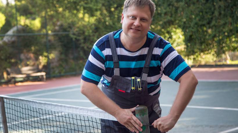 A man wearing a striped, short-sleeved shirt and overalls is repairing the net on his private tennis court.