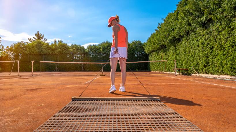 A woman wearing an orange hat, orange tank top, white skirt, and white shoes is sweeping the clay on an outdoor tennis court.