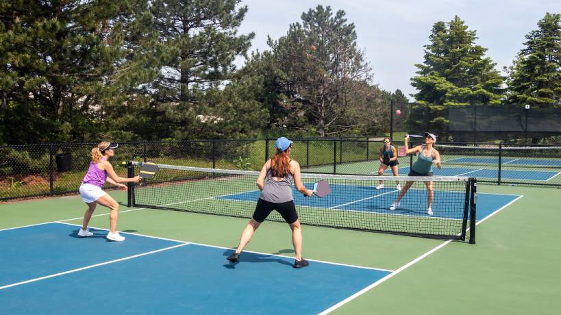 Four women play a doubles pickleball match on outdoor courts surrounded by trees.