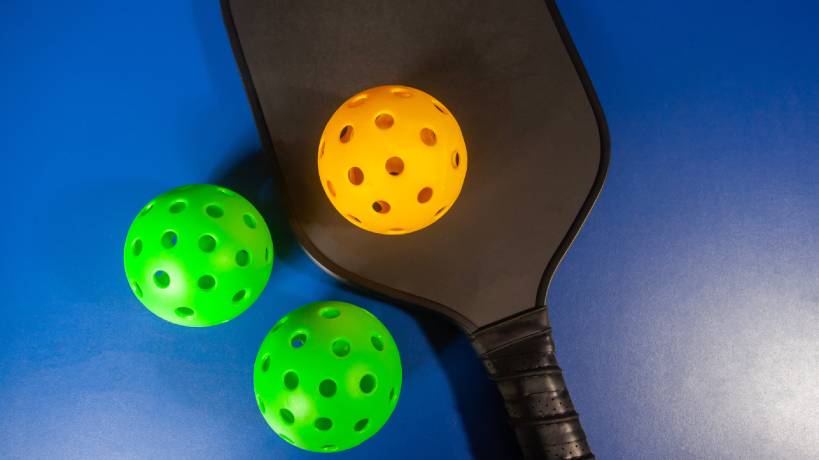 Three pickleball balls, one yellow and two green, and a black pickleball racquet sit against a blue backdrop.