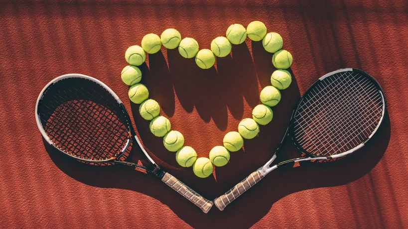 A group of tennis balls is placed in a heart-shaped formation on an orange surface. A pair of racquets surround the tennis balls.