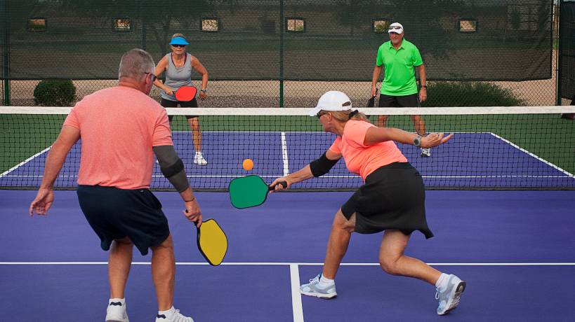 Two mixed doubles teams playing pickleball against one another. The woman on the bottom right hits a backhand shot.