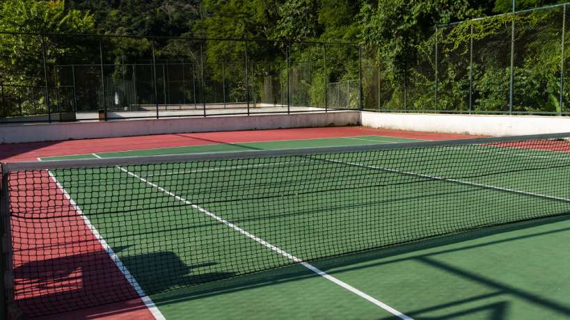 A tennis court with a green playing surface and a brick red perimeter. A natural tree-lined landscape surrounds the court.