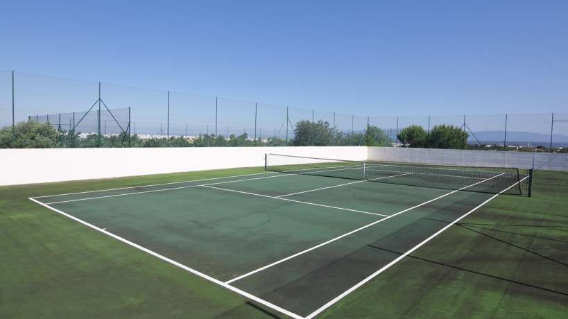 A two-toned tennis court with a sage green playing surface and a moss green perimeter. The open sky surrounds the court.