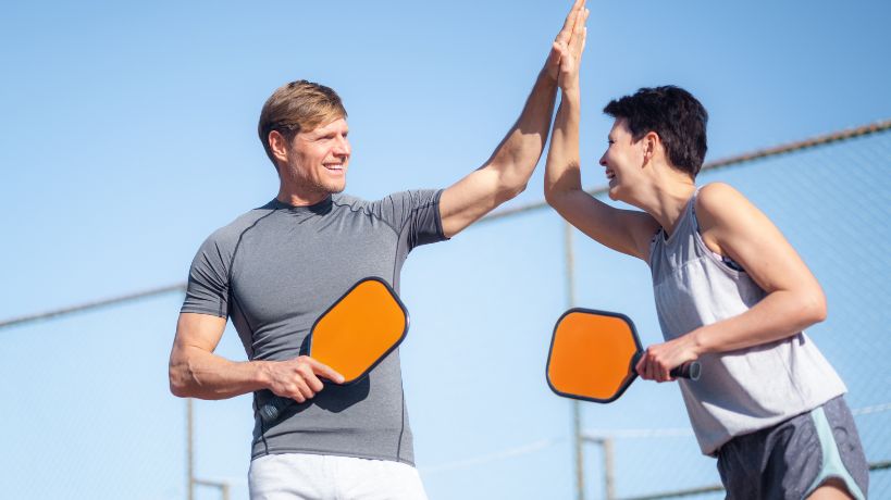 A man and a woman high-five one another while holding orange pickleball racquets. A fence is in the background.