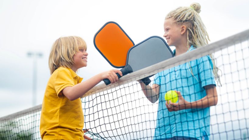 A young boy holding a grey pickleball paddle smiles at a young girl holding an orange pickleball paddle.